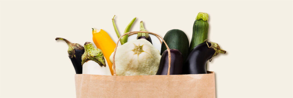 Papiertragetasche gefüllt mit verschiedenen frischen Sommergemüsen wie Zucchini, Auberginen, Patisson und gelber Paprika, vor hellem Hintergrund.